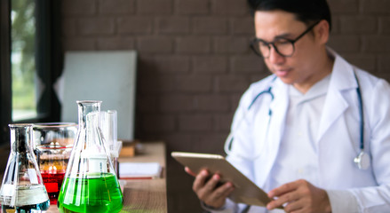 The acting setup of medical work. The selective focued image of the laboratory glassware with the color liquid chemical and the blurred background of young Asian medical doctor reading the reportr.