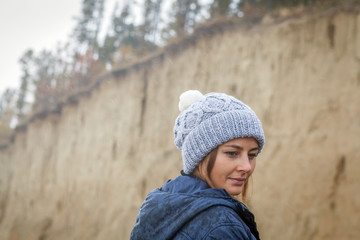 Close-up A young dark-haired woman in a knitted gray cap made of natural wool, a blue jacket on a warm autumn day against the backdrop of a sand pit and coniferous forest
