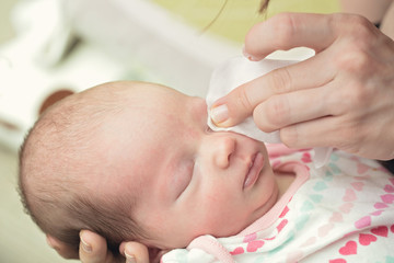 Mother cleaning eyes of a newborn baby with physiological solution on a cotton strip; baby's stork bites visible on eyelid and forehead