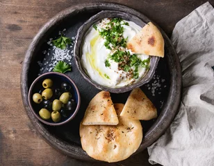  labneh middle eastern lebanese cream cheese dip with olive oil, salt, herbs served with olives, traditional pita bread on terracotta plate over dark texture wooden background. Top view with space © Natasha Breen