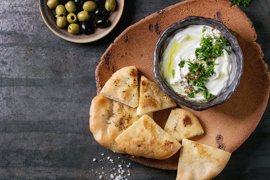 labneh middle eastern lebanese cream cheese dip with olive oil, salt, herbs served with olives, traditional pita bread on terracotta plate over dark texture metal background. Top view with space