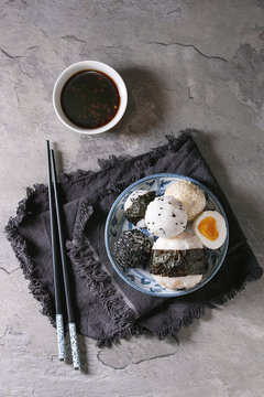 Blue plate with different size rice balls with black sesame and seaweed nori, served with soft boiled eggs, soy sauce, chopsticks over gray table. Asian style dinner. Top view with space