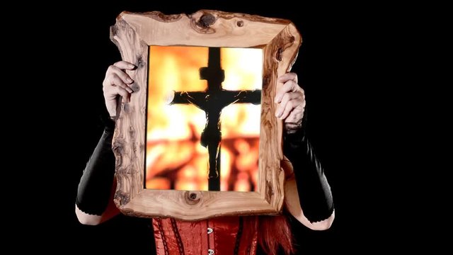 A dark girl holding a wooden frame with a scene: Jesus Christ on the cross (silhouette shadow of a statue) over a fire pyre burning in the night.
