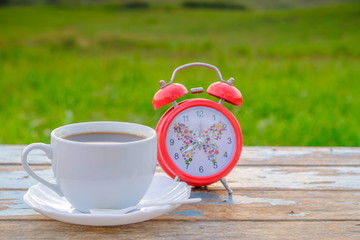 Start up concept . White  coffee cup with red alarm clock on wooden table with nature background