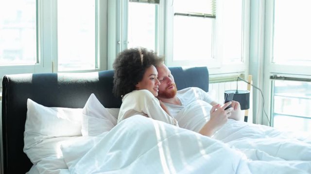 Young couple laughing together and using mobile phone while lying in bed