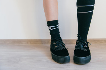 Female feet in black elegant feminine shoes with tankette and high cotton socks standing on wooden floor on white wall background. Unrecognizable kinky odd girl`s legs.