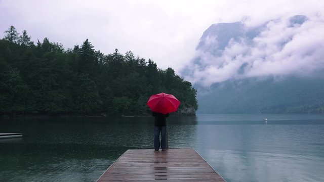 Woman with red umbrella contemplates on rain in front of a lake. Sad and lonely female person looking into distance.