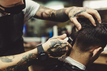 Crop tattooed stylist concentrated on shaving man with machine doing hairstyle.