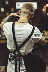 Back view of stylish barber wearing mask and apron and working with client in salon.
