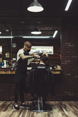 Young tattooed stylist taking care of client in chair doing haircut with clipper.