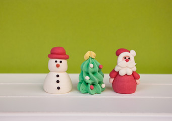 Handmade figures santa claus, new year tree and snowman on white wooden shelf and green background. New year and christmas card.