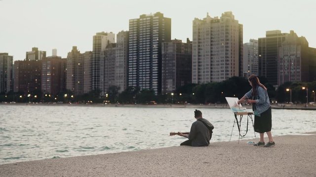 Young Beautiful couple on the shore of Michigan lake, Chicago, America. Woman draw, man play guitar.