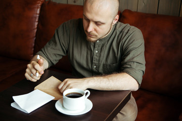 Young serious fashionable man sitting alone in loft-styled cafe and writing a letter. Former factory building, natural daylight.