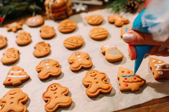 Decoration process of Christmas bakery. Unrecognizable woman decorates homemade gingerbread cookies with blue icing, festive decor around. Family culinary and New Year traditions concept