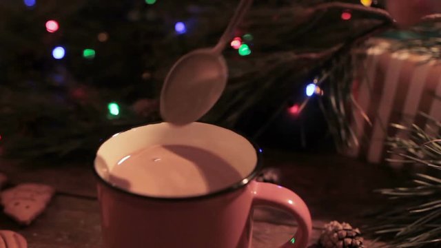 Delicious Christmas holiday with latte. Close up process woman stirring up hot drink and put out the spoon after that. Festive blurred background of illuminated pine on backdrop