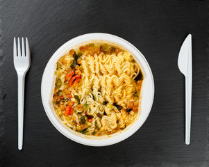 Chinese instant noodles, with red pepper, meat, onions and carrots, in disposable dishes with a fork and knife, on a black stone background.