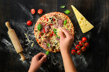 Girl hands making, decopating, preparing pizza with basil leaves on dark background. Top view, copy...