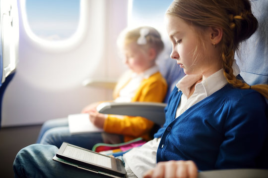 Adorable Little Children Traveling By An Airplane. Girl Sitting By Aircraft Window And Reading Her Ebook During The Flight. Traveling With Kids.