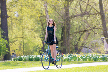 Naklejka premium Portrait view of girl on bicycle wearing on black short dress. Young happy Woman riding along road on green spring outdoor Park. Sporty young girl riding a bicycle on a sunny morning