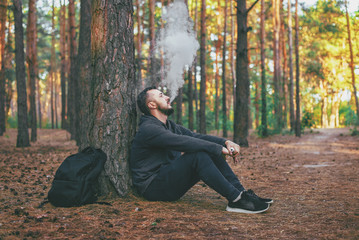 Vape man. brutal bearded young man having rest in forest and vaping an electronic cigarette....