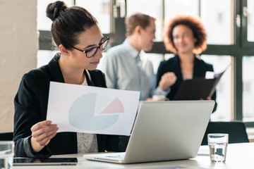 Hard-working young woman analyzing printed and electronic business information while sitting at...