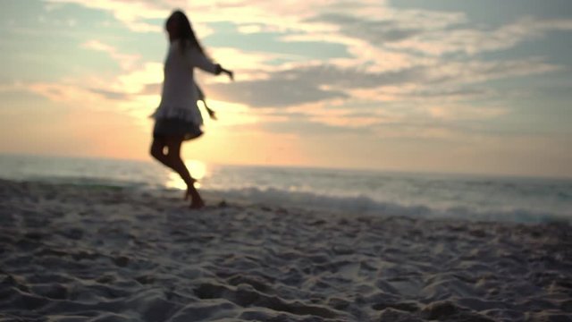 footage in defocus of happy attractive girl with long dark hair dancing spinning with stretched out arms on sand at seashore during wonderful sunset in slow motion