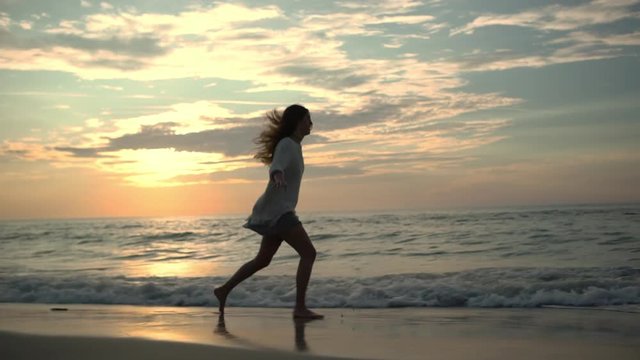 view in profile attractive female person running on wet sand leaving footprints  with stretched out arms along empty beach feeling free like flying while water washing over her feet at picturesque dus