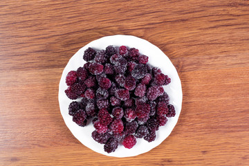 Plate with black sweet blackberry covered with ice on a wooden background. Stocking up for winter from frozen berries.