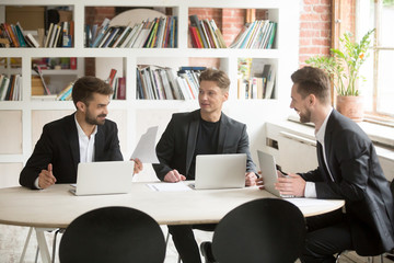 Smiling coworkers discuss positive business matters in front of laptops during briefing. Executives talking about optimistic company goals. Good corporate news, telling jokes, being funny concept.