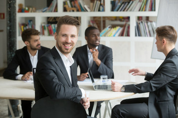 Handsome young smiling businessman looks at camera during business meeting. Team of male corporate colleagues on background, Friendly male executive, successful company representative, CEO or manager.
