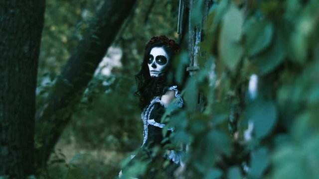 The Mexican Day of the Dead. The young woman with spooky make-up for Halloween dressed in black clothes leaning to wooden wall and looking at camera. Shooting through tree branches. 4K