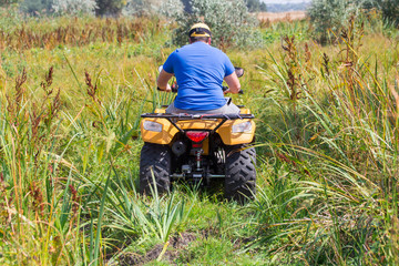 Caucasian man in sport protective goggles riding an ATV quad bike over rough terrain between high grass bushes. Adventure activity concept