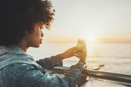 Close-up side view of beautiful young African American lady with curly afro hair, leaning her hands on chrome railing and shooting dramatic sunset over the sea, using back camera of her smartphone