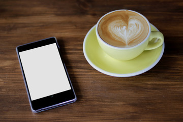 Coffee latte art heart shape in light green ceramic cup besides white smart phone with white black screen on wooden table background,mock up image 