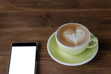 Fototapeta na wymiar Coffee latte art heart shape in light green ceramic cup besides white smart phone with white black screen on wooden table background,mock up image 