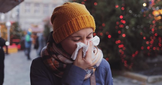 Sick Woman Blowing Her Nose Into Tissue Outdoors. SLOW MOTION 4K DCi. Young Female having cold or flu symptoms, standing in a busy city street. Autumn or winter cold weather.