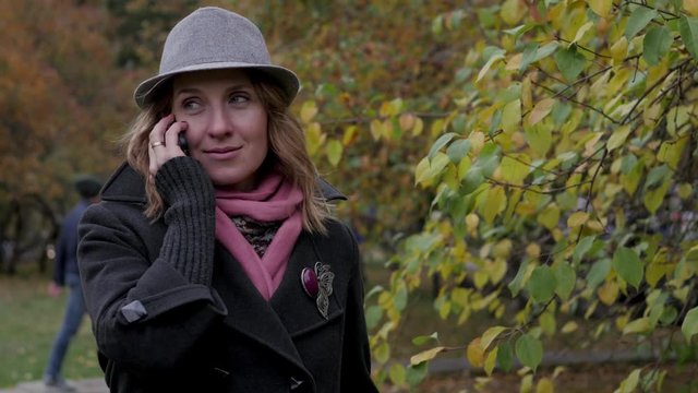 Beautiful young woman talking on a mobile phone in an autumn park wearing a hat