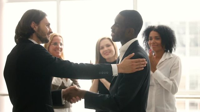 Boss promoting rewarding african american male worker, appreciating for good job, businessmen wearing suit congratulating shaking hands with applauding staff standing in office, employee recognition