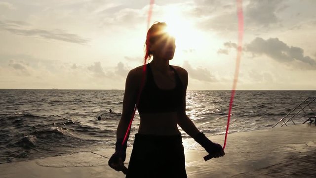 Closup view of a young woman working out on the jump rope against the sun by the beach in slowmotion. Girl jumping on a skipping rope by the sea