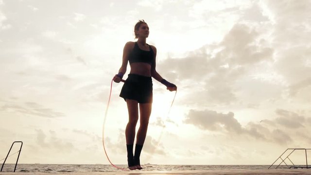Athletic woman in shorts and sport bra working out on the jump rope against the sun by the beach in slowmotion. Girl jumping on a skipping rope by the sea