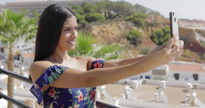 Excited young girl in colorful dress using smartphone and posing while taking selfie on background of tropical resort area.