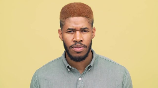 closeup dark-skinned guy with stylish haircut in blue skirt looking down then looking at camera and smiling broadly with white teeth over yellow background. Concept of emotions