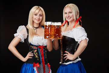 Two beautiful blonde women are holding glasses of beer in hands and stand on black background in studio.
