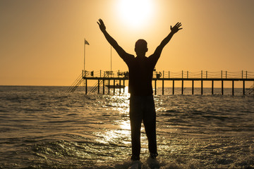 Fototapeta premium Man standing on the beach, raising his hands above his head at the sunset