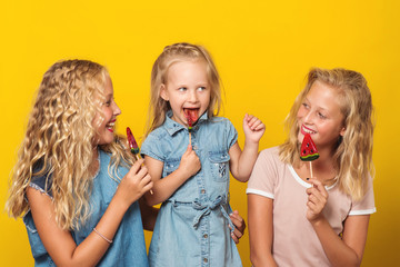 Happy children eating fruit lollipops in studio, cute girls sisters or friends having fun together isolated over yellow background.