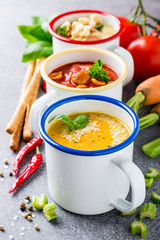 Assorted delicious homemade soups in enamel mugs with ingredients. Healthy food concept.