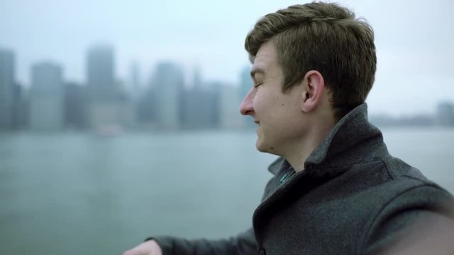 Young man holding camera in front of his face and waving his hand and pointing to Manhattan island