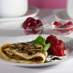 Serving of homemade crepes with strawberries, chocolate topping and powdered sugar. Small jars of red berries and teacup in background.
