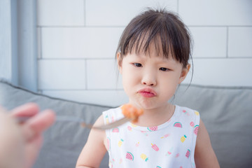 Little asian girl with unhappy face between mother want her to have lunch,picky eater child. - obrazy, fototapety, plakaty