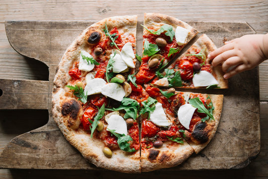 Sliced Pizza On Chopping Board, Child's Hand Reaching For Pizza Slice, Overhead View